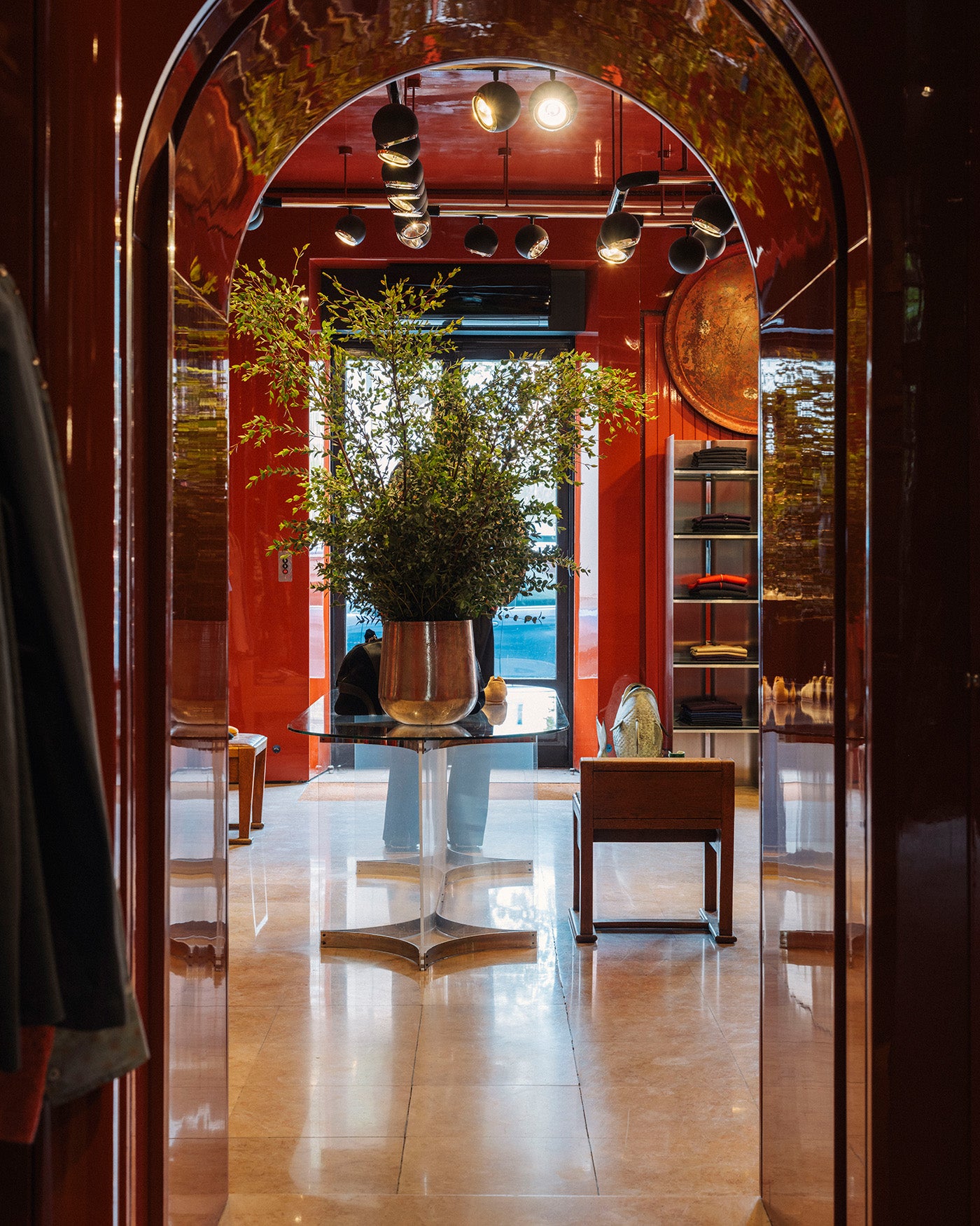 A view through a dark arched doorway into a modern interior with warm red walls, polished beige floors, a glass-top table with a large green plant, and shelving.