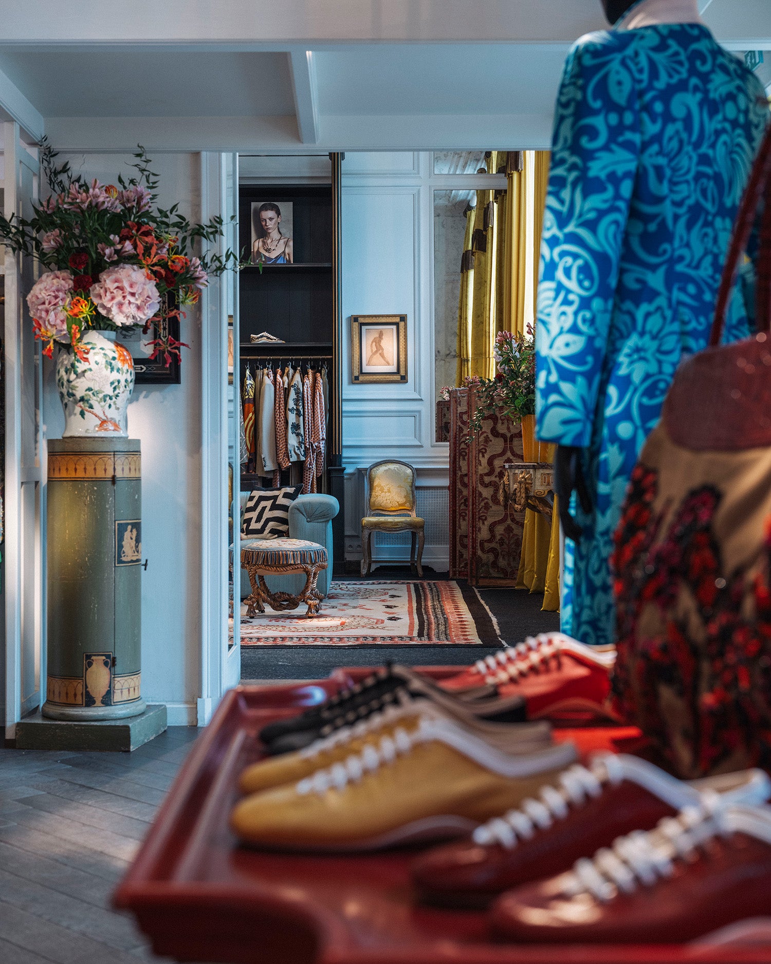 A styled interior scene showing a mannequin wearing a bright blue ornate patterned coat, a red table of shoes with the foreground and a boutique walkway in the background.