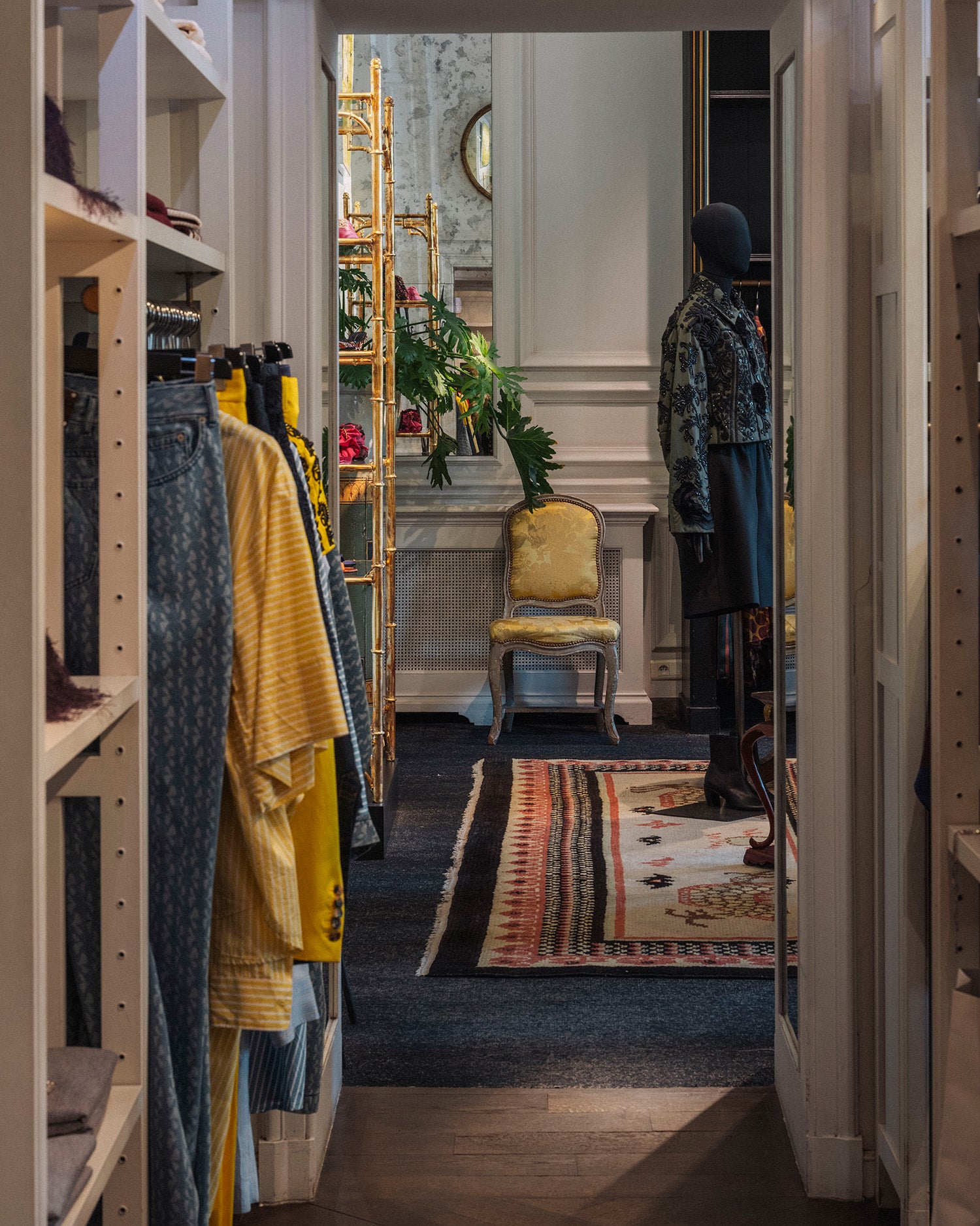 A boutique interior hallway with clothing racks on the left, a mannequin wearing a dark patterned outfit on the right, and a yellow upholstered chair near a mirror in the background.