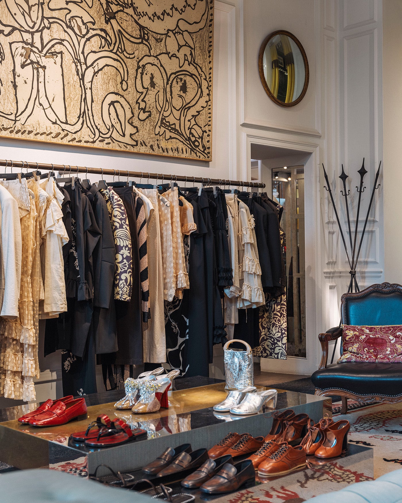 A luxury boutique interior showcasing a collection of fashion items, including a clothing rack with patterned garments, a blue velvet armchair with a patterned cushion, and multiple pairs of shoes displayed on a glass table.