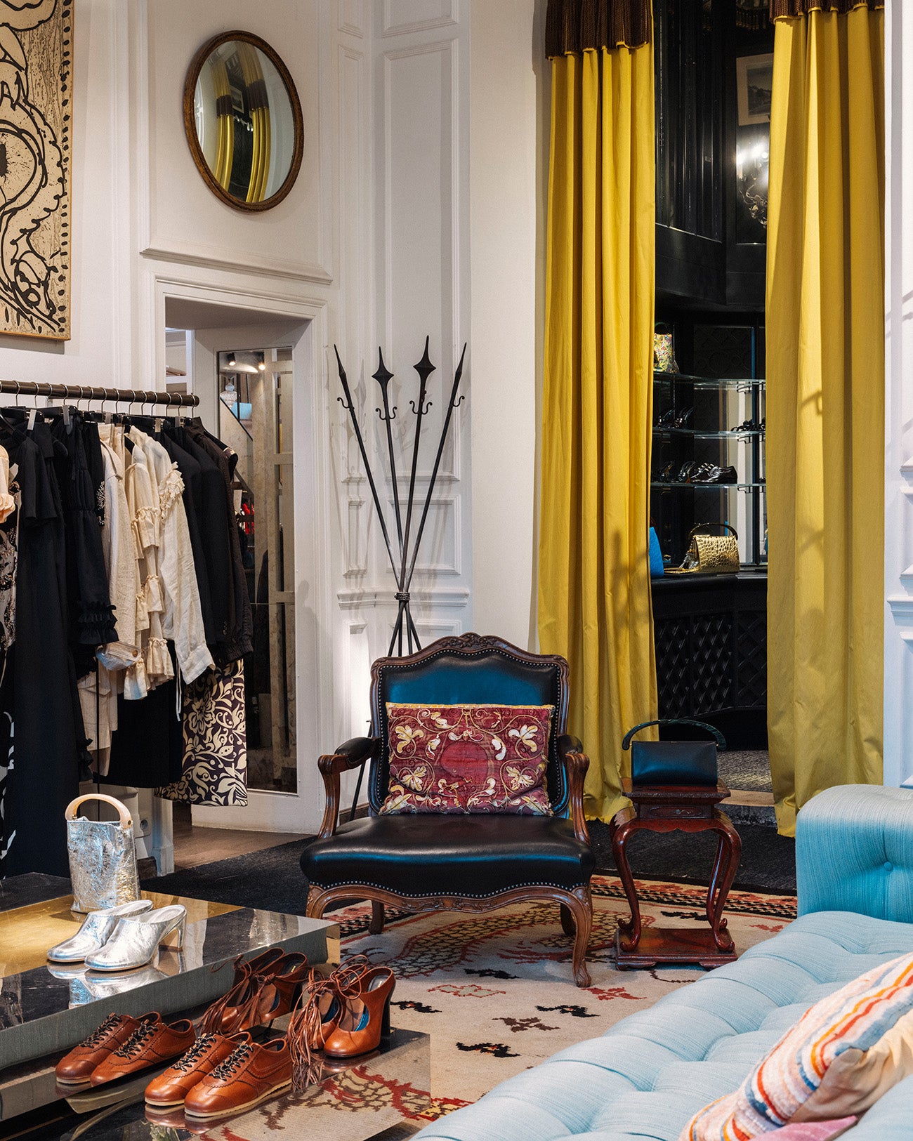 A styled interior scene showing a blue upholstered armchair with a patterned red and gold pillow, mustard yellow curtains, a light blue sofa, brown leather shoes on a glass table, and clothing racks in a boutique room.