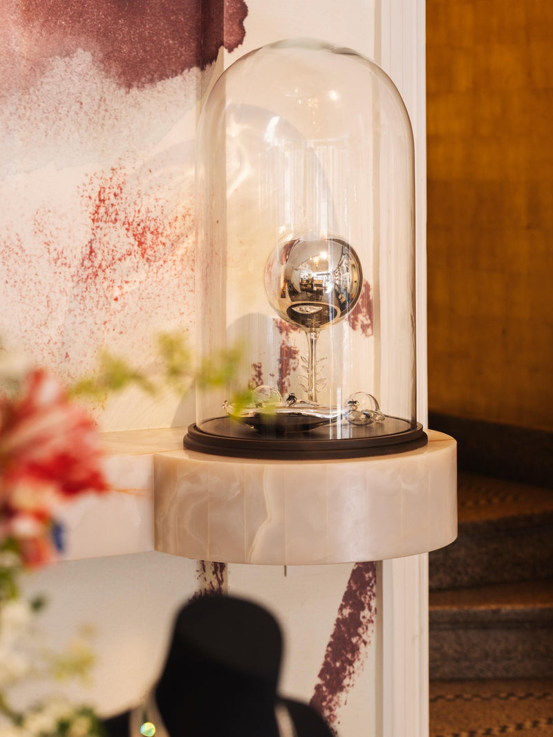 Decorative art piece with glass cover on a dark brown base against a painted background and staircase.