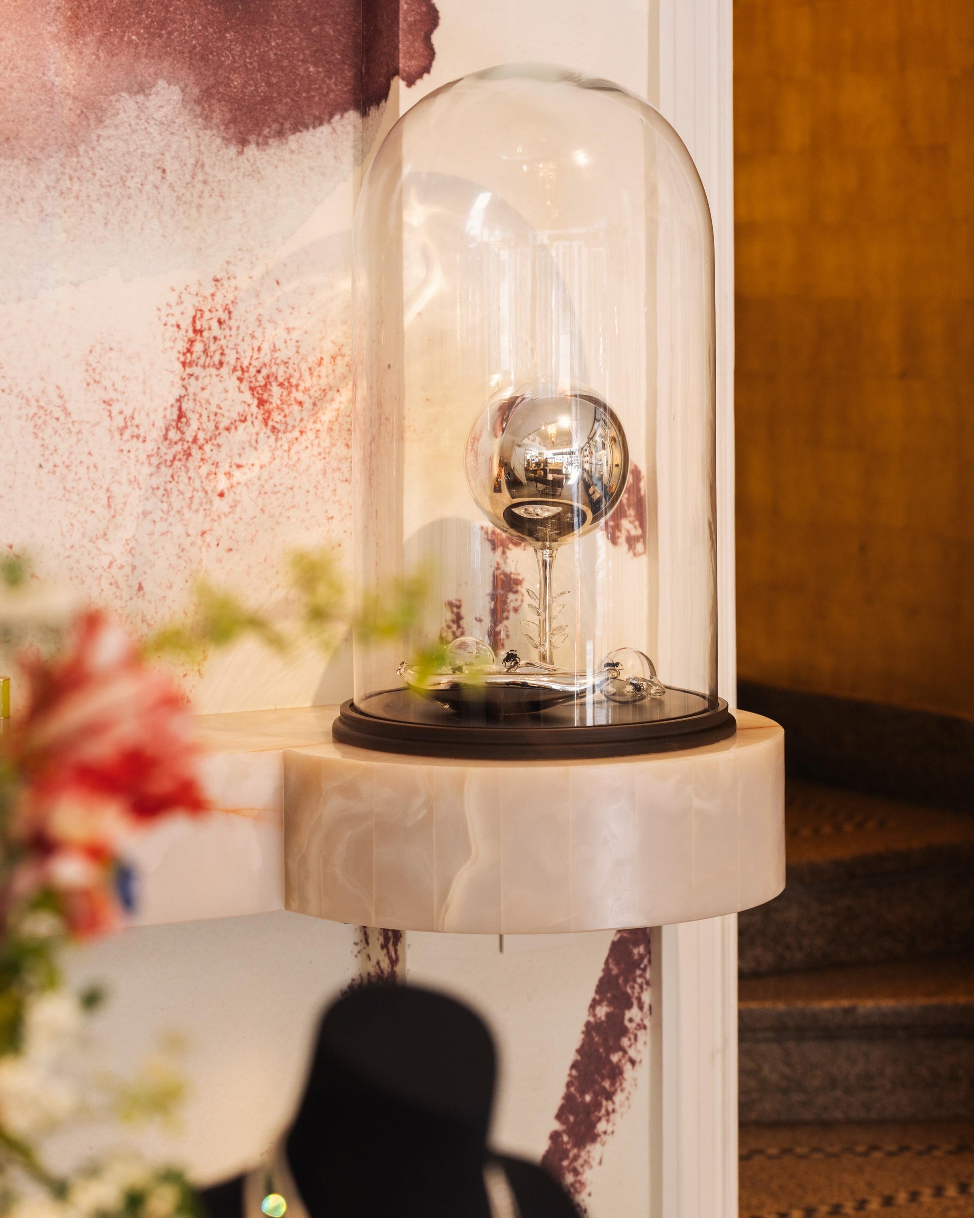 Decorative art piece with glass cover on a dark brown base against a painted background and staircase.