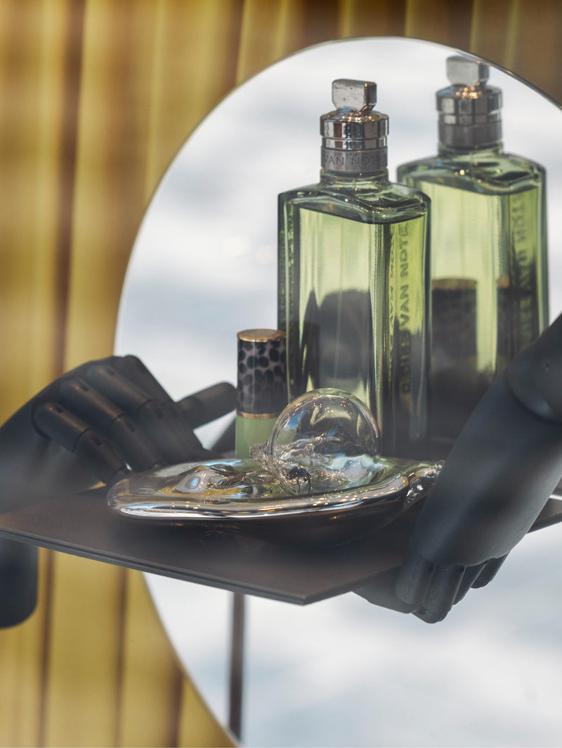 A green Mystic Moss eau de toilette bottle alongside a lipstick case and glass object, on a tray held by two black wooden hands in front of a circular mirror