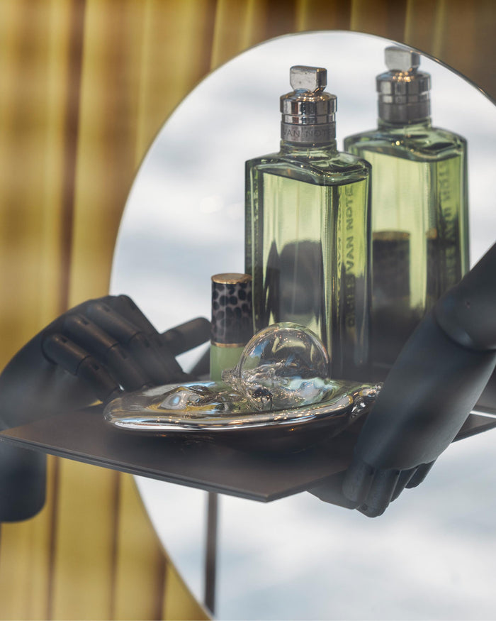 A green Mystic Moss eau de toilette bottle alongside a lipstick case and glass object, on a tray held by two black wooden hands in front of a circular mirror
