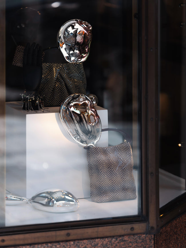 A window display featuring two Dries Van Noten handbags amidst three metallic sculptures, mirroring their surroundings.