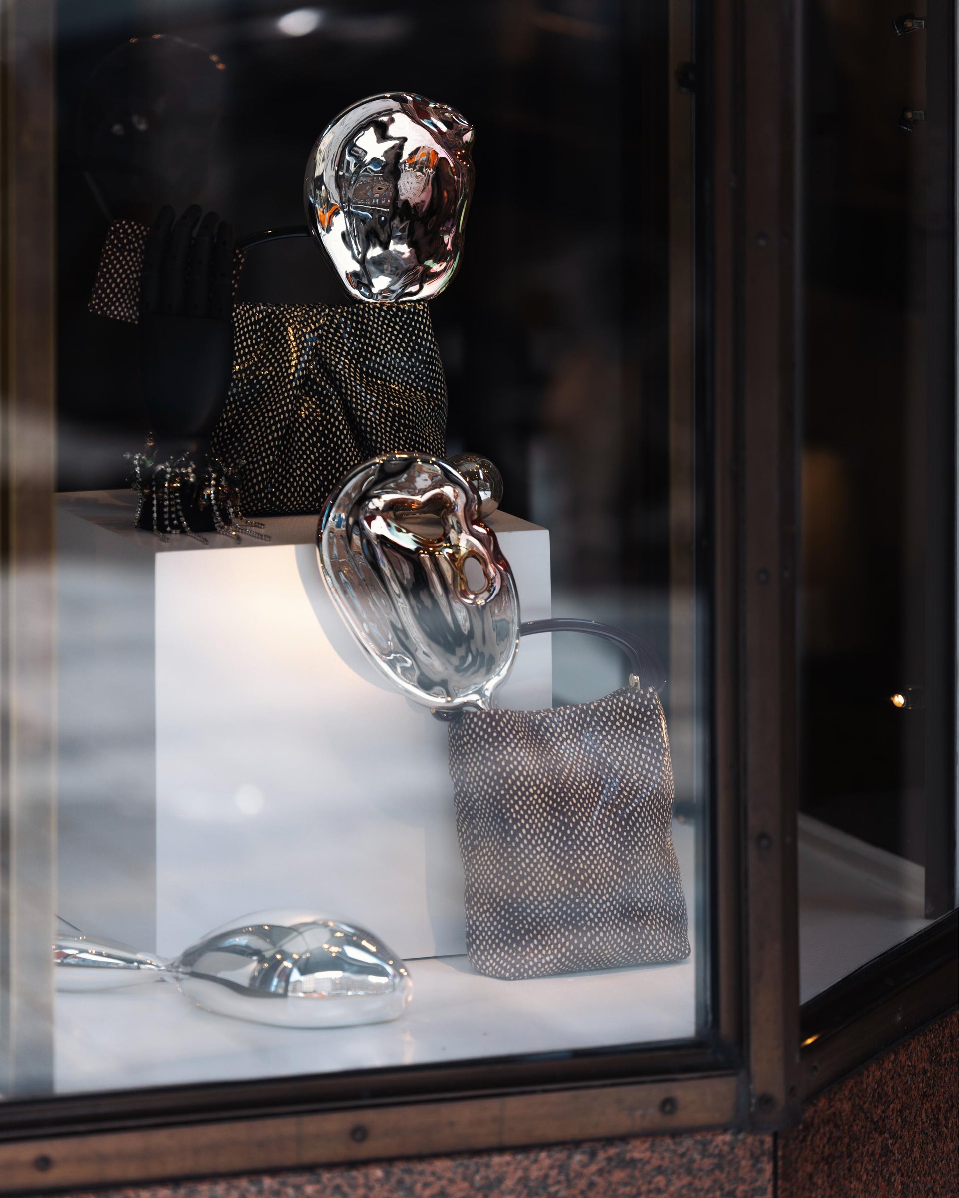 A window display featuring two Dries Van Noten handbags amidst three metallic sculptures, mirroring their surroundings.