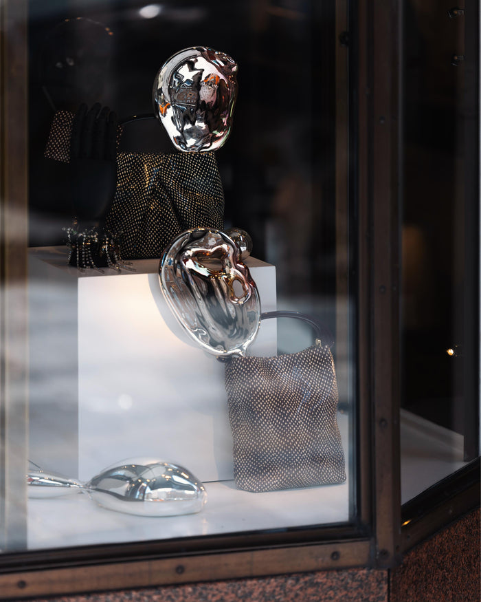A window display featuring two Dries Van Noten handbags amidst three metallic sculptures, mirroring their surroundings.