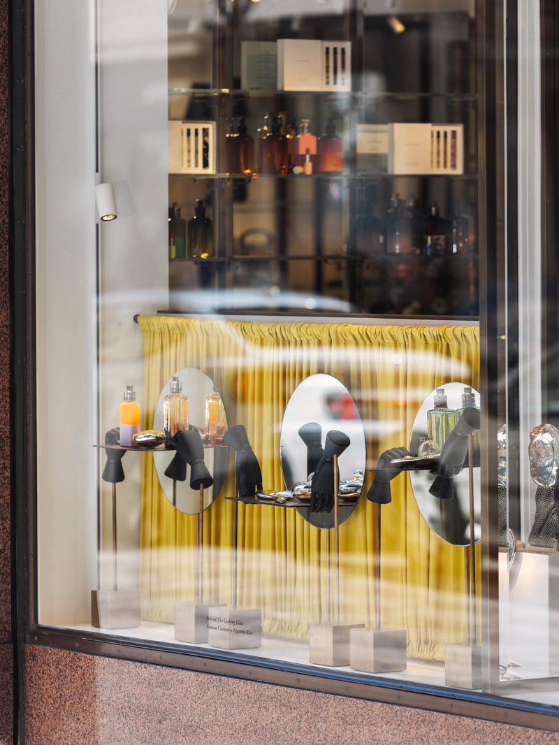Display of perfume bottles amidst small art sculptures, in a store window with mirrors and a yellow curtain behind them.