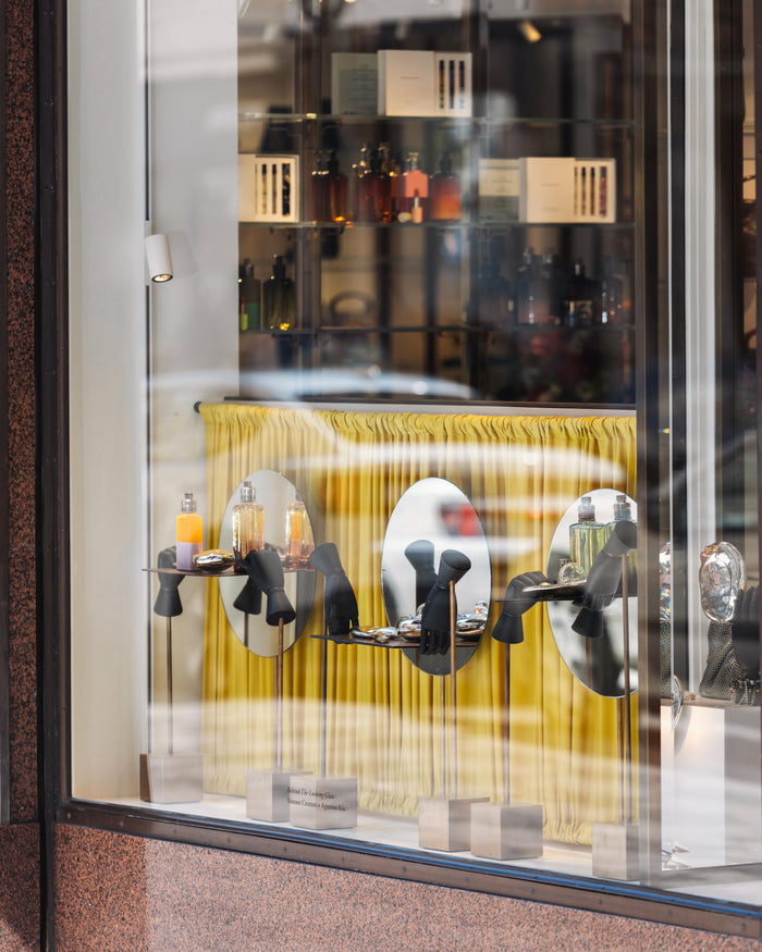 Display of perfume bottles amidst small art sculptures, in a store window with mirrors and a yellow curtain behind them.