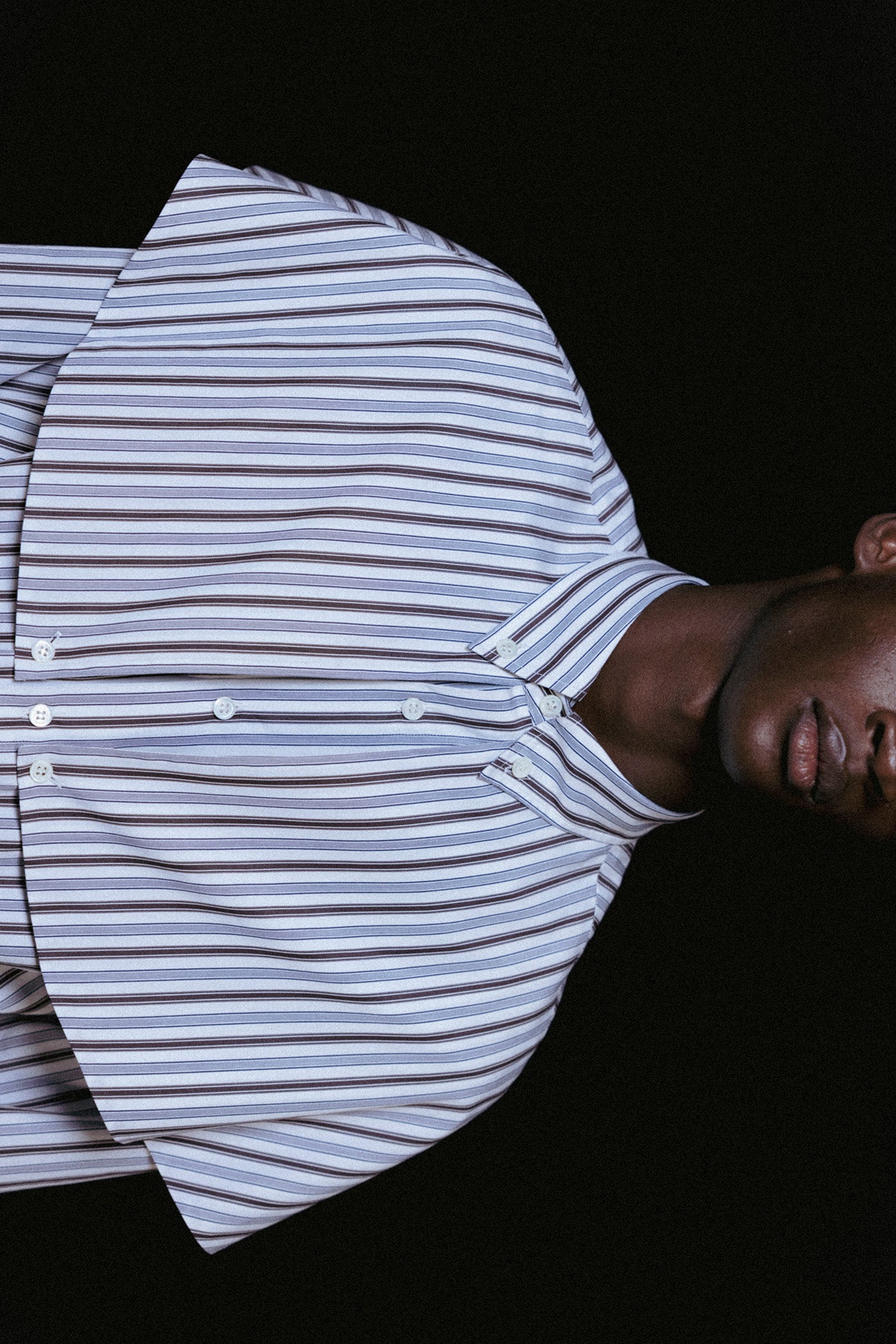 A close-up side view of a person wearing a white button-up shirt with blue and gray horizontal stripes, photographed against a black background.