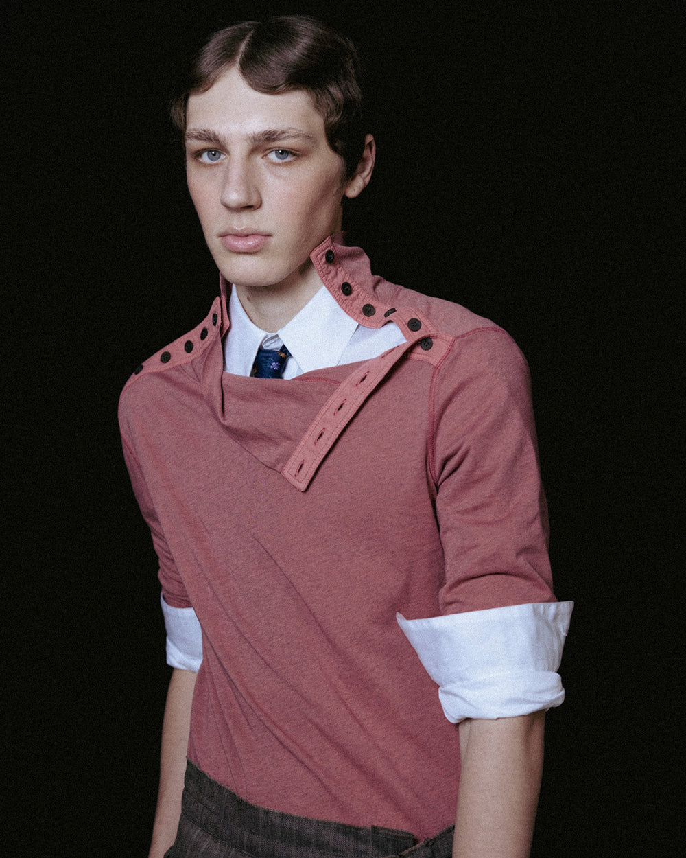 A model wearing a unique dusty rose long-sleeve top with structured shoulder details, layered over a white collared shirt and a blue patterned tie, set against a solid black background.