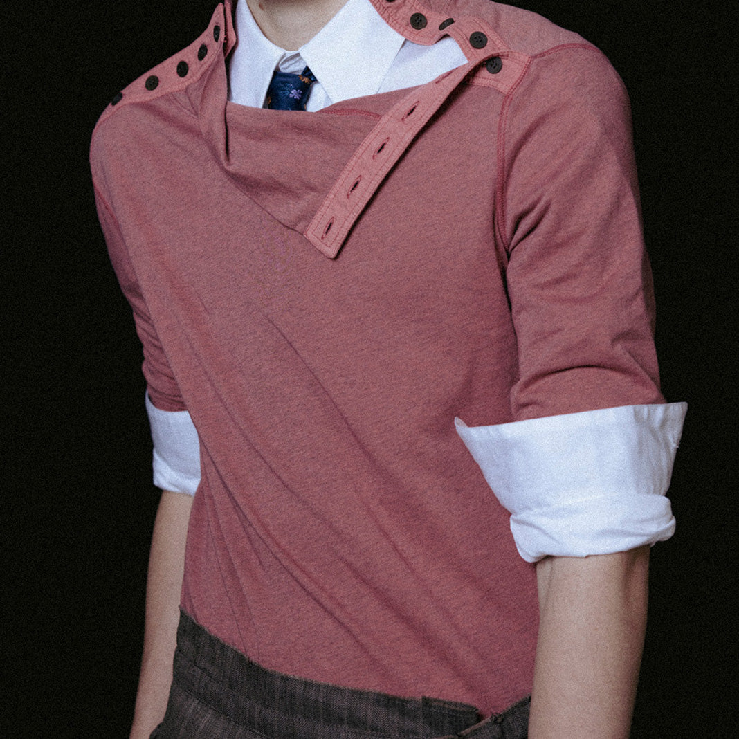 A model wearing a unique dusty rose long-sleeve top with structured shoulder details, layered over a white collared shirt and a blue patterned tie, set against a solid black background.