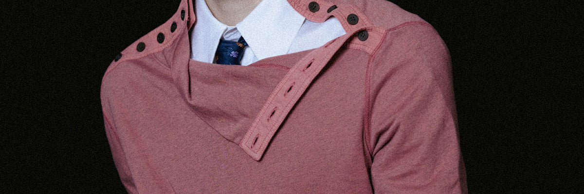 Close-up of a model wearing a dusty pink draped top with an asymmetric buttoned collar over a white shirt and floral tie, against a black background.