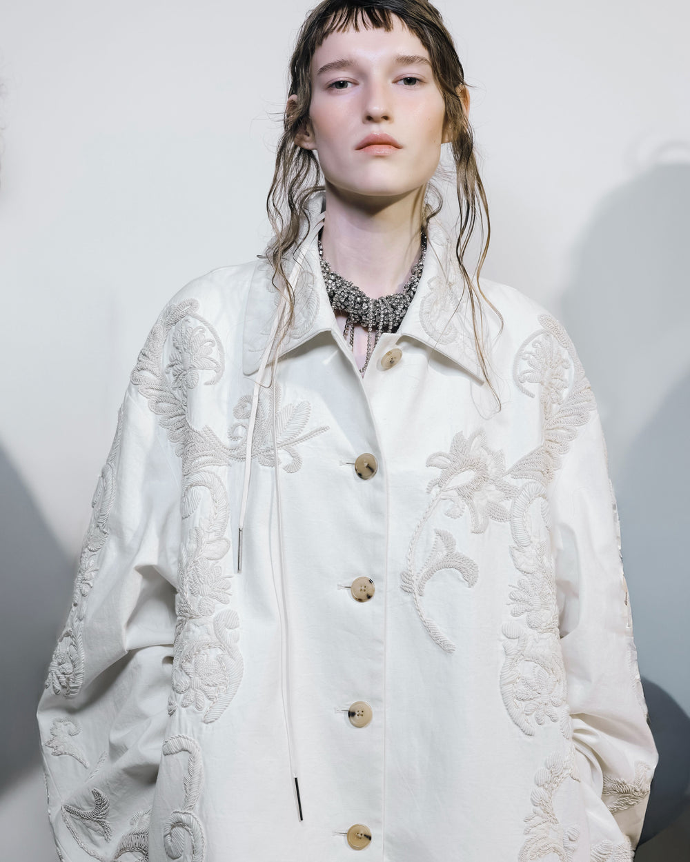 Person wearing a white embroidered coat and metallic necklace against a neutral background