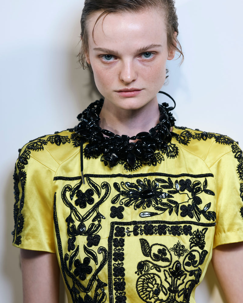 Model wearing a yellow top with black embroidery against a plain background