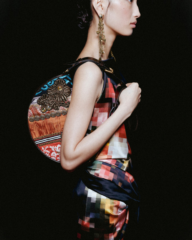 Model in a multicolored check dress carrying an embellished patchwork shoulder bag and wearing a long gold-tone earring.