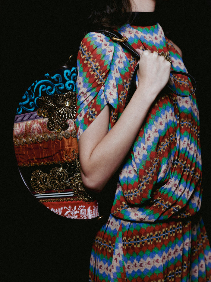 Person wearing a colorful patterned dress holding a decorative handbag against a dark background