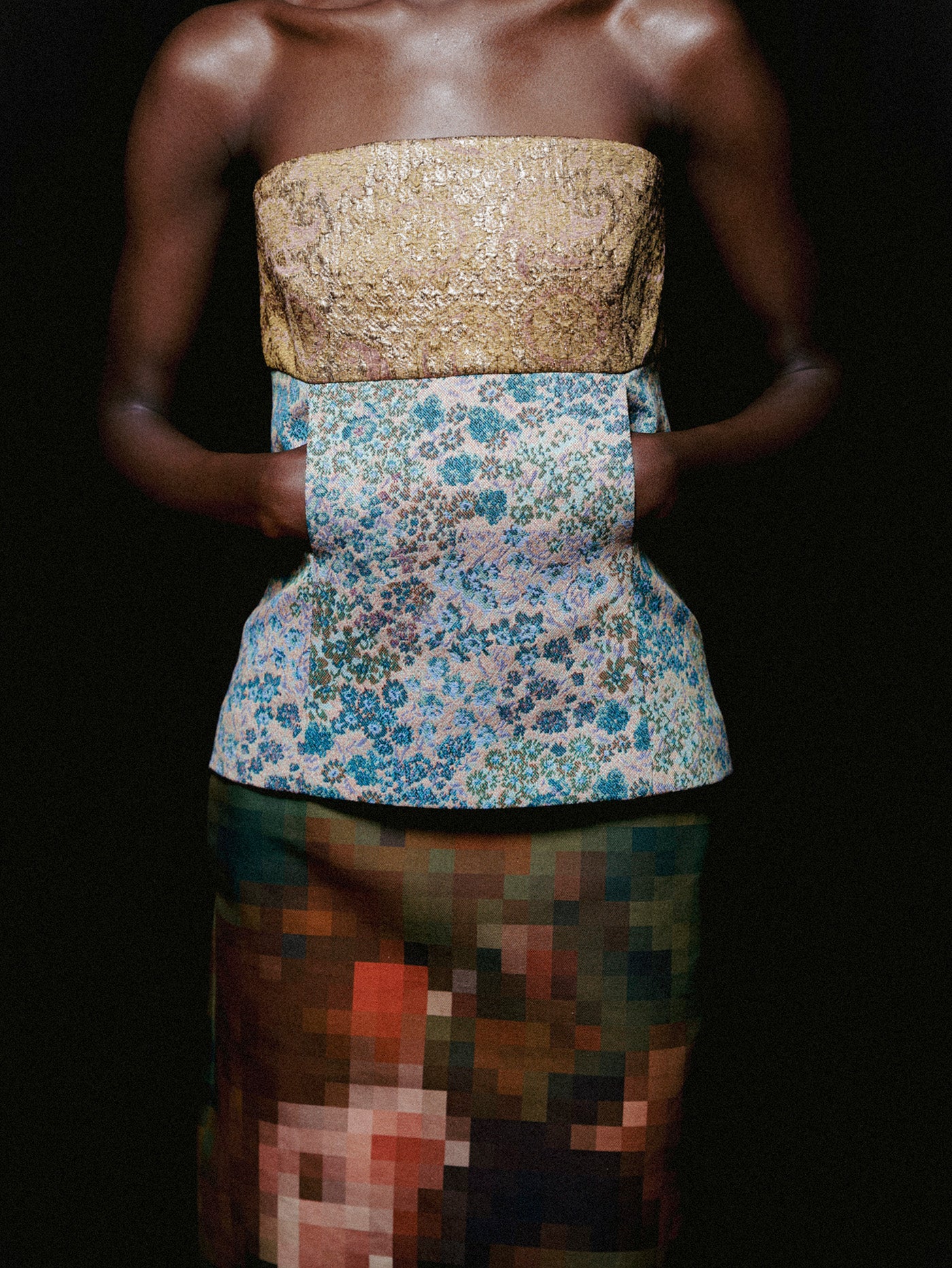 A model is shown from the shoulders down wearing a strapless top with a textured gold upper section and a blue floral patterned lower section, paired with a pixelated skirt against a black background.