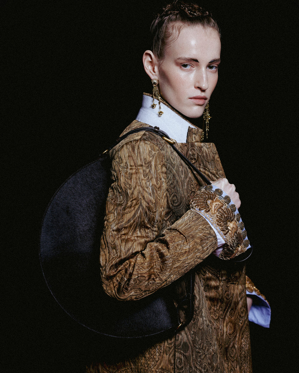 Model wearing a textured brown coat, shirt with embellished cuffs and long gold earrings, carrying a large round black shoulder bag against a dark background.