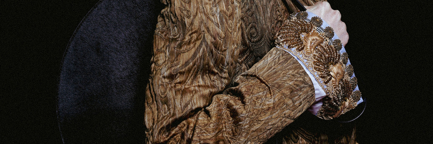 Close-up of a model wearing a textured brown coat with an ornate embellished cuff, carrying a large round black shoulder bag against a dark background.