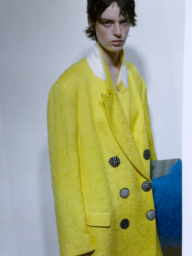 Person wearing a bright yellow textured coat with black buttons on a plain background